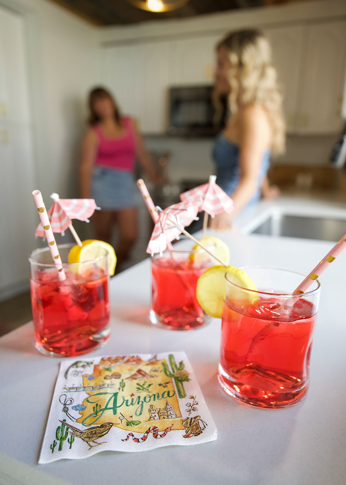 Three drinks with straws and umbrellas on a table, a woman in the background, and a tablecloth with 'Arizona' written on it.
