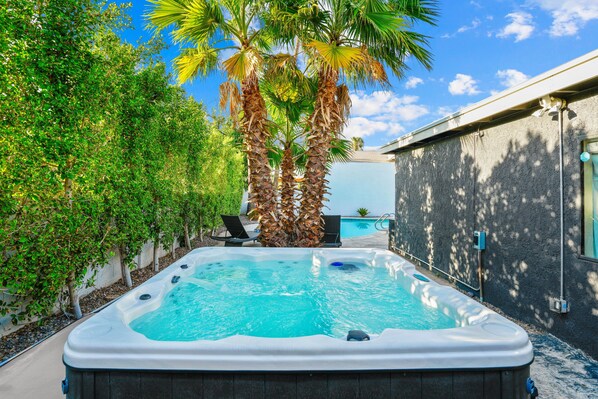 Un jacuzzi dans un jardin, avec une piscine, entouré de palmiers et d’une maison.