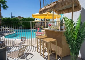 Un espace bar aménagé au bord de la piscine, avec des meubles en osier, un flotteur en forme de flamant rose et un parasol à toit de chaume.