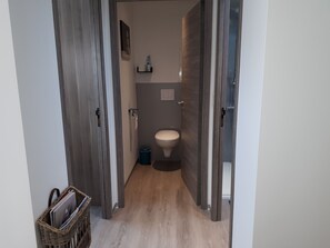 A narrow hallway leading to a bathroom with a toilet, a small trash bin, and a shelf with toiletries.