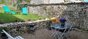 A patio area with a stone wall, a grill, a table with chairs, and two blue lounge chairs.