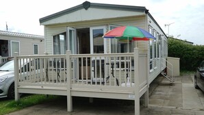 A mobile home with a fenced porch, a car parked nearby, and a colorful umbrella. - Cozy Hunstanton Caravan Sleeps 8 Pets go Free (Hunstanton)