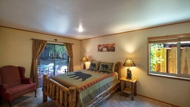A bedroom with a large bed, two bedside lamps, a chair, a window with a view of the outdoors, and a painting on the wall. - Scenic Wonders Apple Tree Home 3 Br (Yosemite National Park)