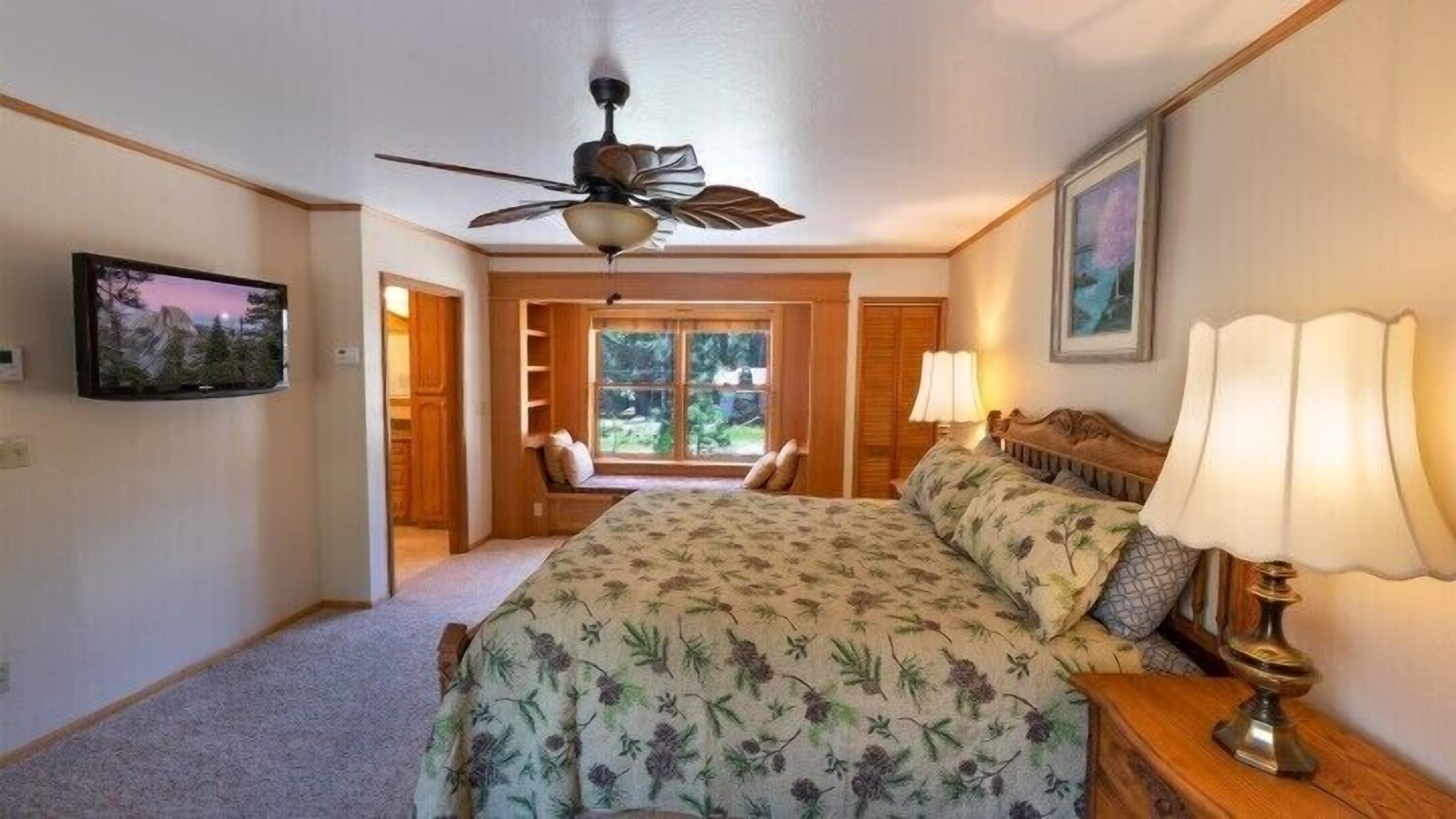 A bedroom with a bed, a ceiling fan, a window with a view of greenery, a bedside table with a lamp, and a framed picture on the wall.