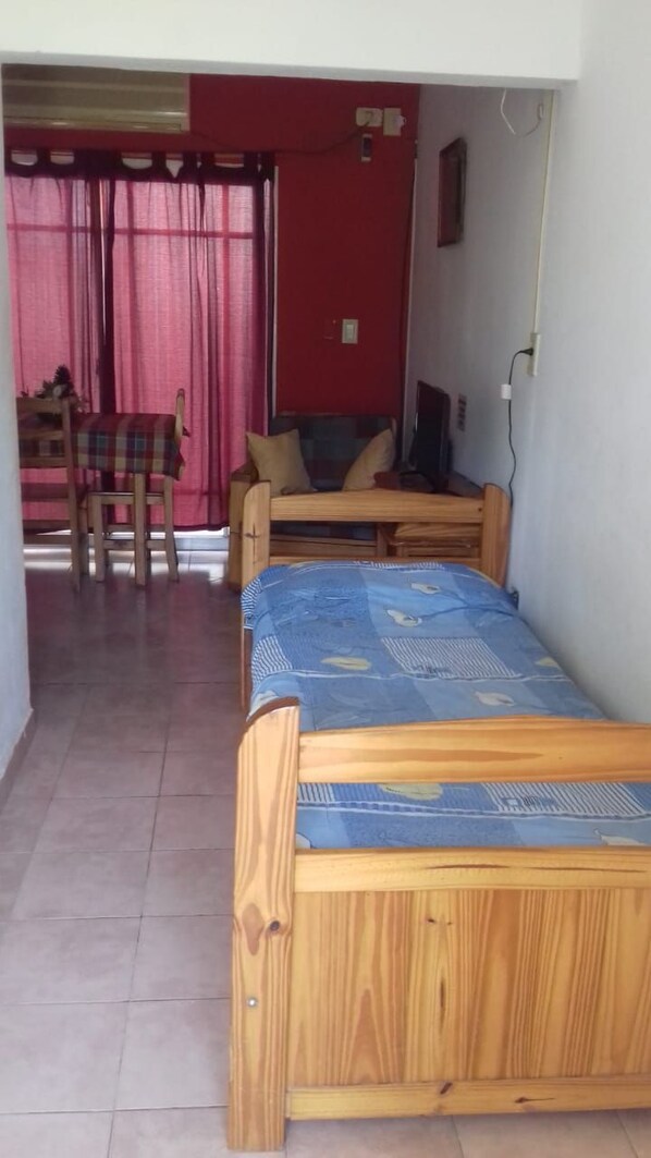 Una habitación con dos camas de madera, una pared roja, una ventana con cortinas y una pequeña mesa con una silla.