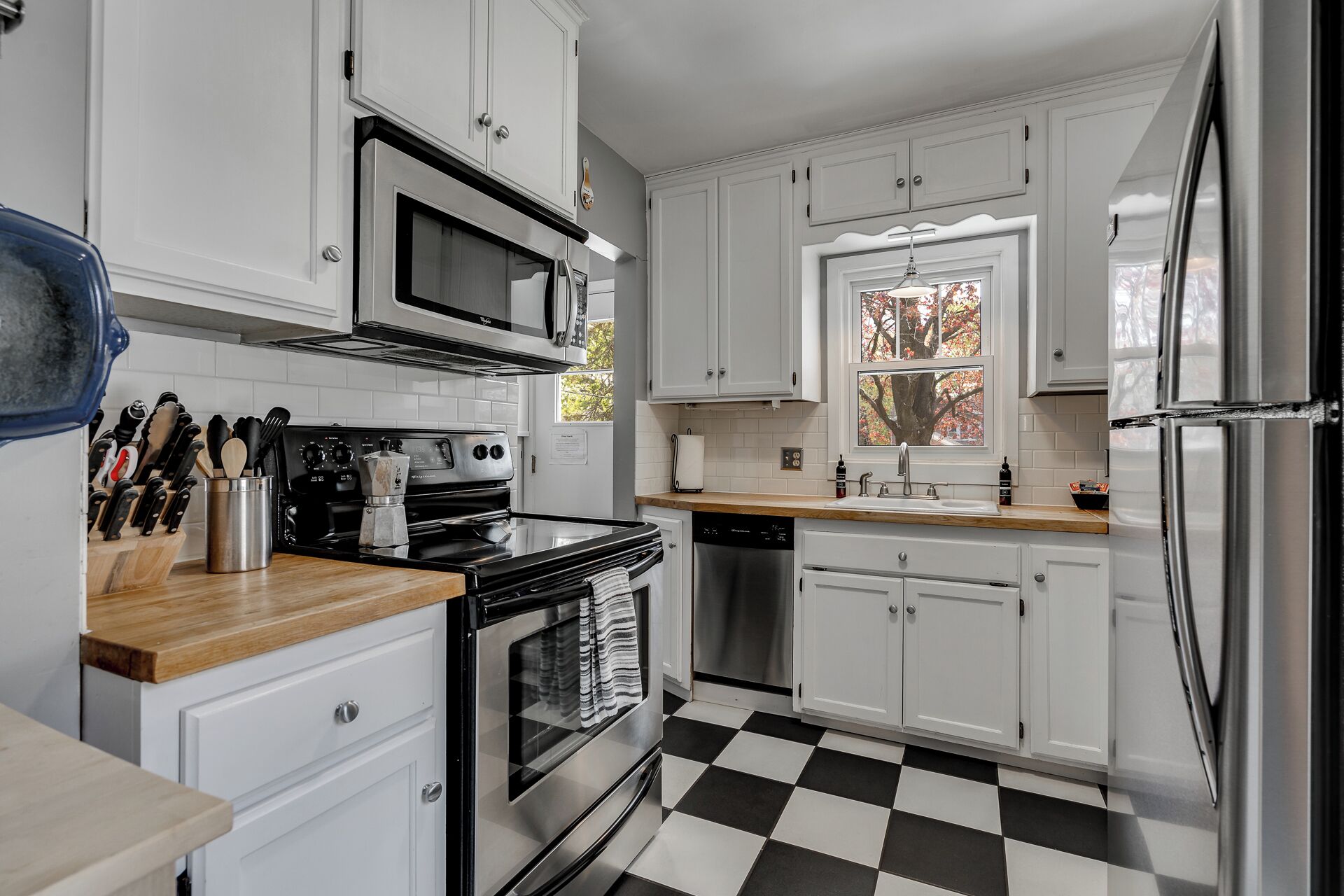 A kitchen with white cabinets, a black and white checkered floor, stainless steel appliances, and a window with autumn leaves visible outside.