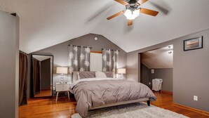 Quarto com teto inclinado, cama com tapete branco, duas mesas de cabeceira com luminárias e um ventilador de teto.