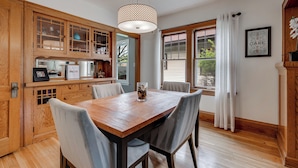 Uma área de refeições com mesa e cadeiras de madeira, luminária pendente e vista para o exterior através de uma janela com cortinas.