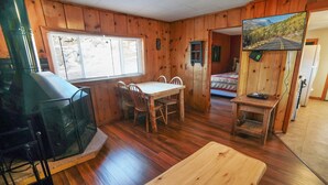 A cabin interior with a wooden dining table, chairs, a TV, and a painting of a mountain road. - Charming Two bedroom Cabin, Dog Friendly close to National Park (Estes Park)
