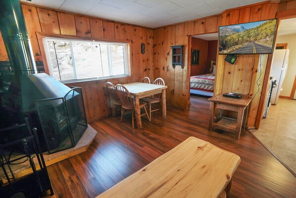 A cabin interior with a wooden dining table, chairs, a TV, and a painting of a mountain road. - Charming Two bedroom Cabin, Dog Friendly close to National Park (Estes Park)