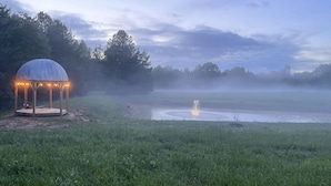 Um gazebo com luzes de festa em um campo gramado com um lago e névoa.
