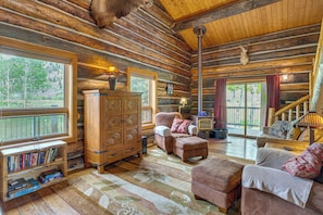 A log cabin interior with a fireplace, wooden furniture, a bookshelf, and large windows.