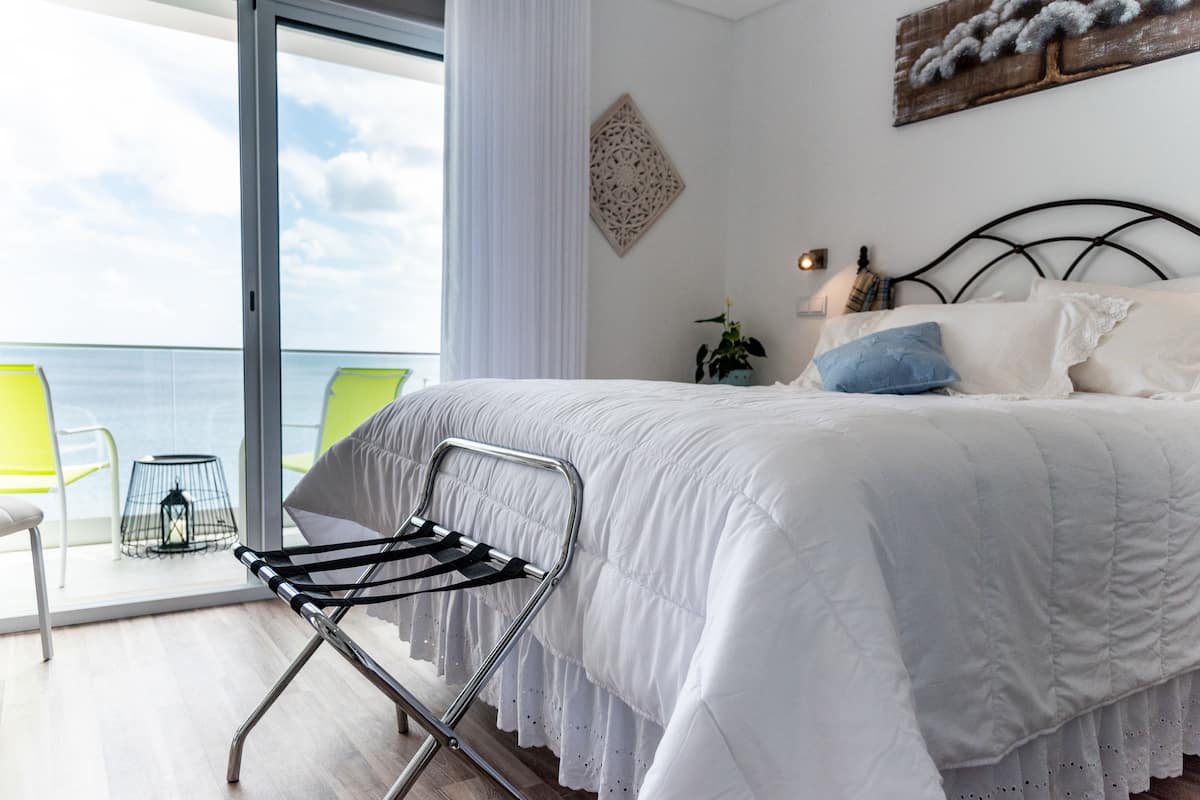 A bedroom with a bed, a chair, a plant, and a view of the sea.