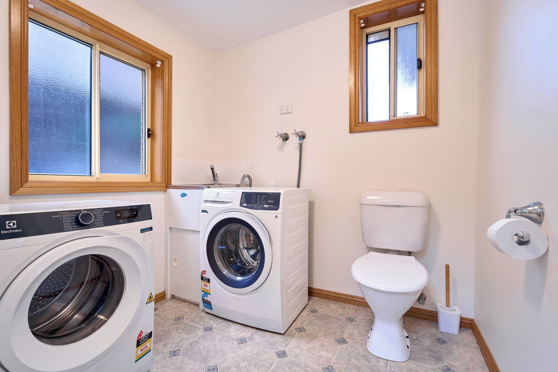 A laundry room with a front-loading washing machine, a dryer, a toilet, and a window with a wooden frame.