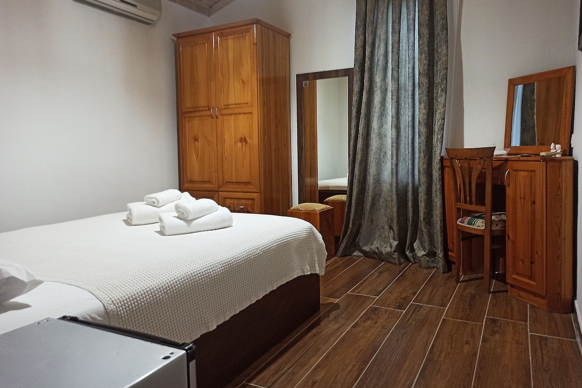 A bedroom with a bed, wooden wardrobe, a desk with a chair, a mirror, and a window with curtains.