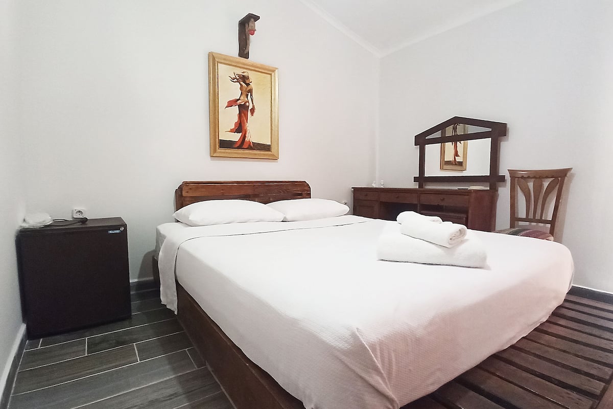 A neatly made bed with white linens, a wooden headboard, a small bedside table, a nightstand, a framed picture on the wall, and a wooden chair.