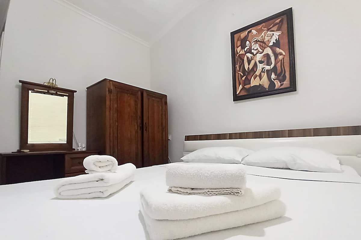 A hotel room with a bed, white linens, a wooden wardrobe, a mirror, and a painting on the wall.