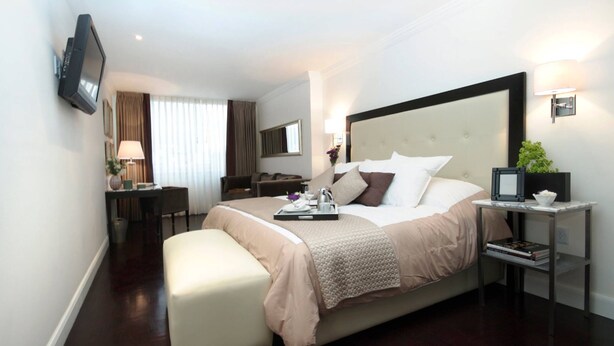 A modern hotel room with a large bed, a bedside table with a lamp, a TV mounted on the wall, and a small desk with a chair. - Suites Londres 212 (Mexico City)