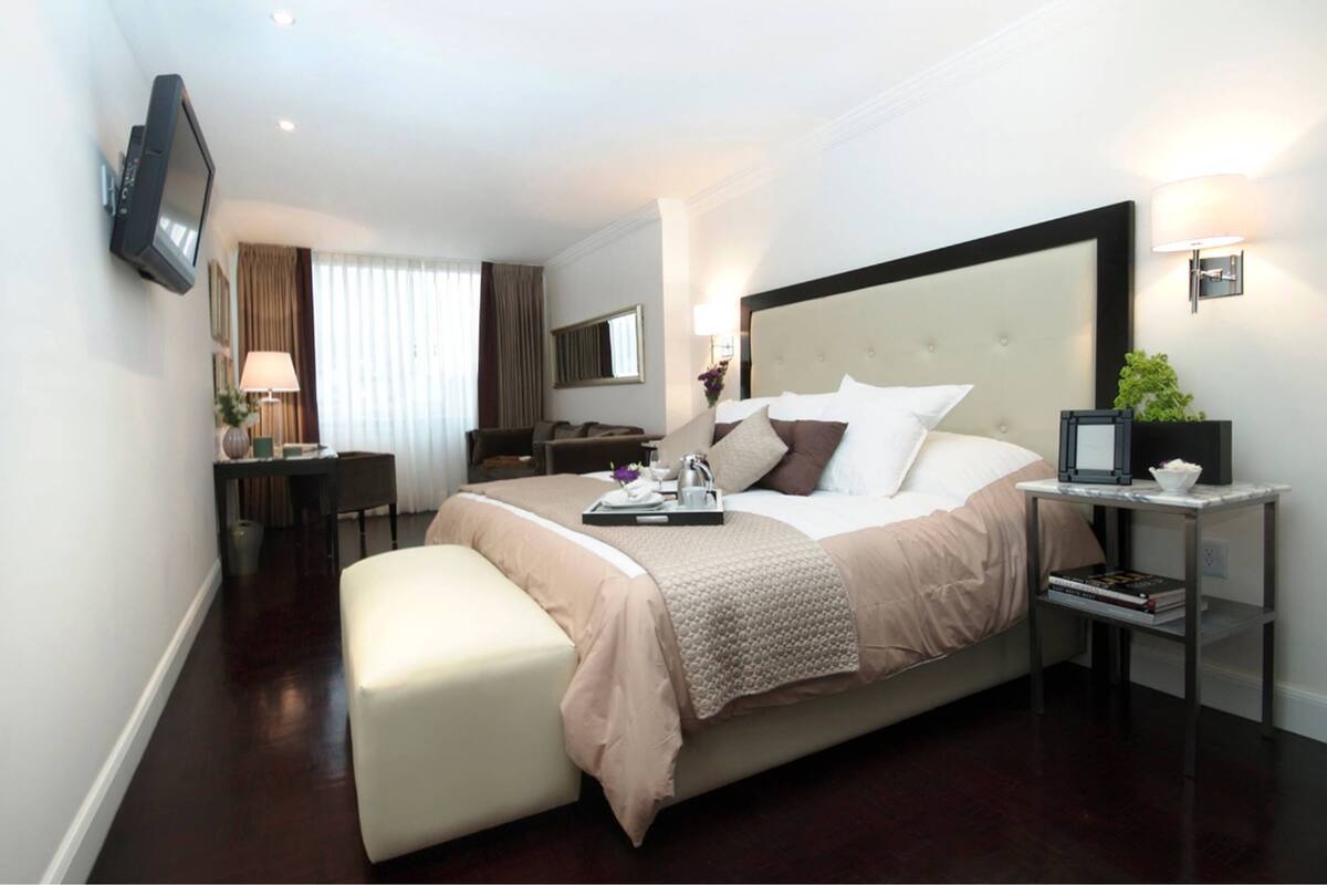 Una habitación de hotel moderna con una cama grande, una mesita de noche con lámpara, un televisor montado en la pared y un escritorio pequeño con silla.