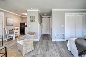 A modern, well-lit room with a wicker chair, a bed, and a TV. A door leads to another room, and there's a sign on the wall. - Walk to Surfside Beach: Studio w/ Covered Balcony (Surfside Beach)