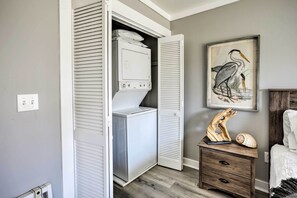 A laundry room with a washer and dryer, a wooden nightstand with a shell and a wooden sculpture, and a painting of a bird on the wall. - Walk to Surfside Beach: Studio w/ Covered Balcony (Surfside Beach)