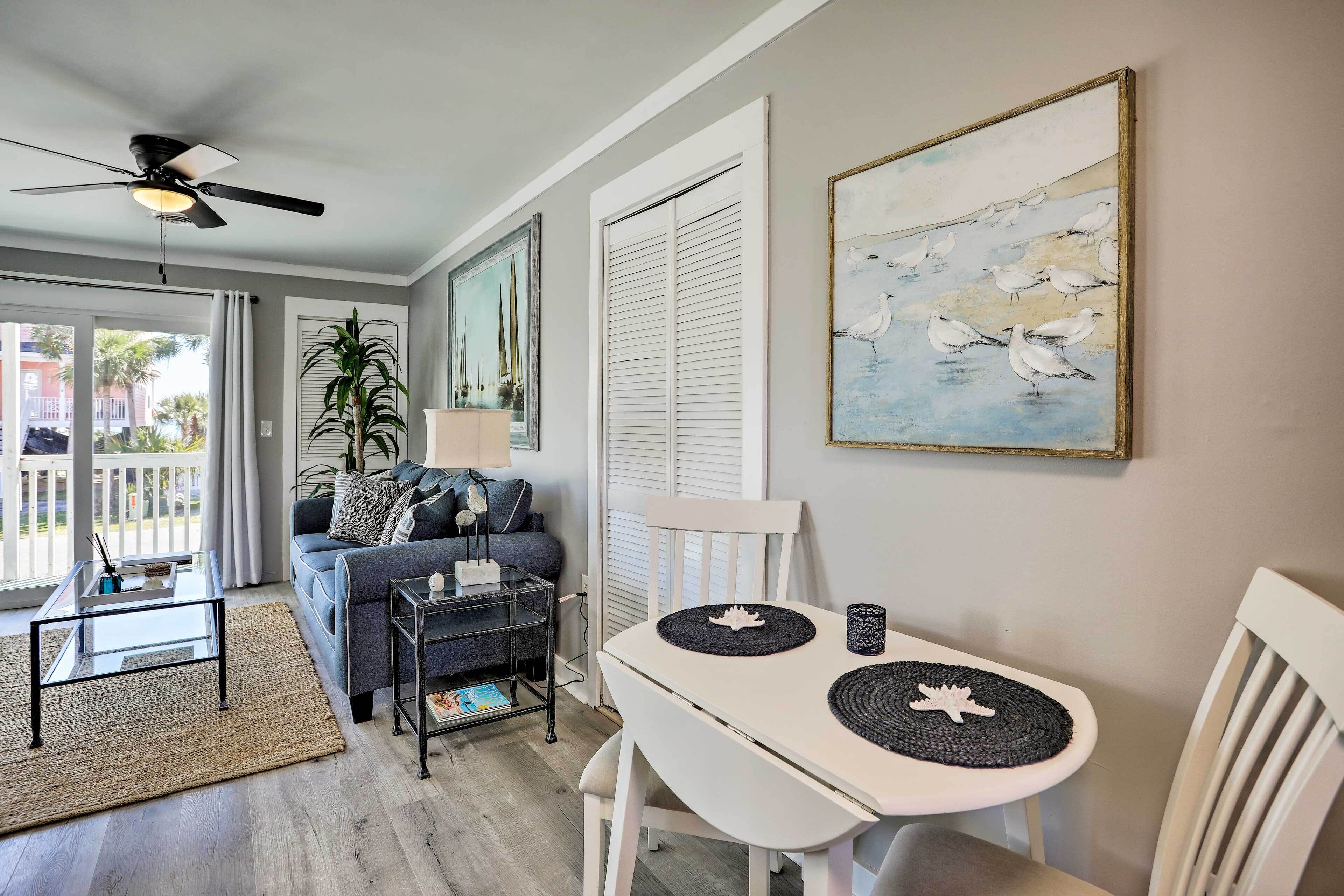 A living room with a white sofa, a glass coffee table, a white dining table with placemats, a ceiling fan, and a painting of seagulls.