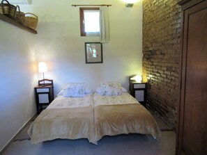 Una cama doble con ropa de cama floral, dos mesitas de noche con lámparas, una ventana pequeña con cortina y una pared de ladrillo.