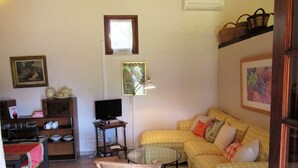A living room with a yellow checkered sofa, a wicker chair, a glass coffee table, a television on a wooden stand, a bookshelf, and a window with a lace curtain. - Vivelorural: Alpendre de Ayala, piscina y jardín (Santa Brígida)