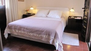 Una cama bien hecha con colcha blanca y lavanda, dos lámparas de noche, una mesita con un florero y una puerta con pomo.