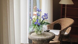 Una mesa de mimbre con un florero y dos copas de vino, una silla de mimbre, y una lámpara en una habitación con ventana y cortinas.