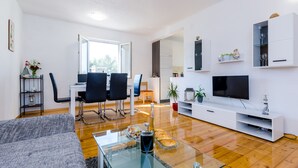 A modern living room with a glass coffee table, a sofa, a dining area with chairs, a TV, and a white entertainment unit. - Apartment & Rooms Maždin - One Bedroom Apartment with Balcony and Sea View (Slano)