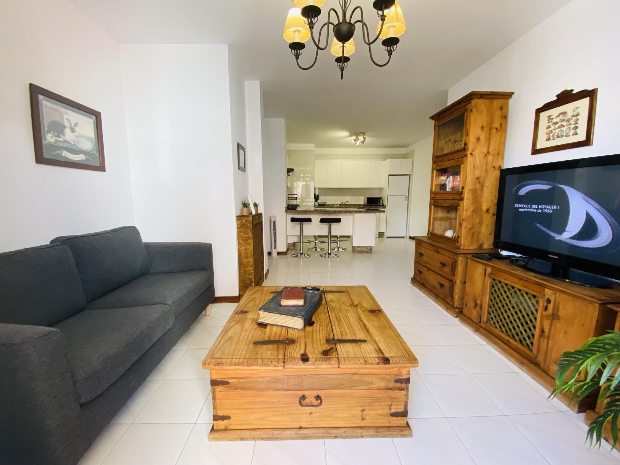 A living room with a sofa, a wooden coffee table with books, a TV, and a kitchen in the background.