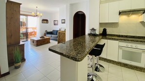 Eine moderne Küche mit weißen Schränken, einer Granit-Arbeitsplatte und einem eingebauten Backofen. Es gibt einen Essbereich mit einem Holztisch und Stühlen sowie ein Wohnzimmer mit einer Couch und einem Couchtisch.