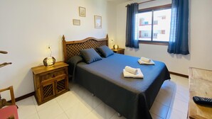 Ein Schlafzimmer mit einem großen Bett, zwei Nachttischen, einer Ablage, einem Fenster mit Vorhängen und einer Deckenleuchte.
