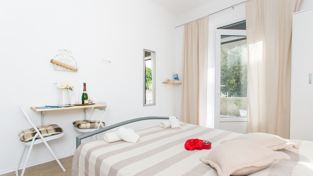 A modern hotel room with a bed, a desk, a chair, a window with curtains, and a view of greenery outside. - Studios IM - Studio Apartment with Shared Terrace (A1) (Dubrovnik)