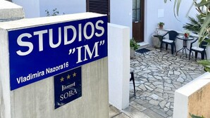 A sign for "STUDIOS 'IM'" with a name and address, a stone-tiled courtyard, outdoor seating, and a white building with shutters. - Studios IM - Studio Apartment with Shared Terrace (A1) (Dubrovnik)