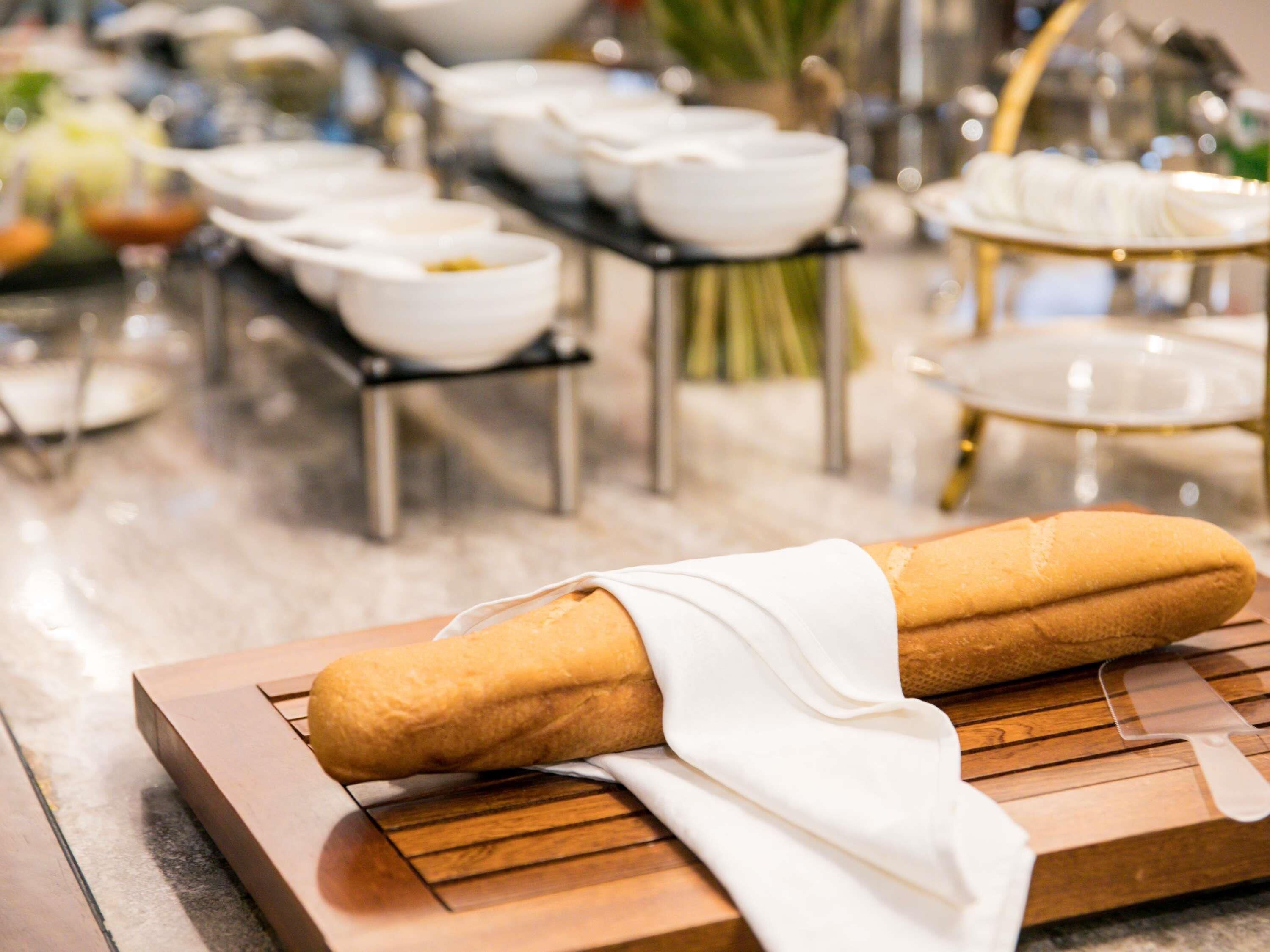 A baguette on a wooden board, surrounded by white bowls and a glass pitcher on a table.
