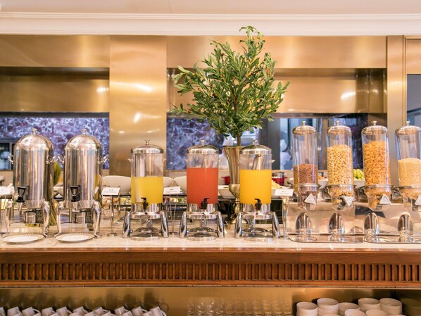 Mostrador de buffet con diferentes dispensadores de comida, incluyendo frutas, cereales y jugos.
