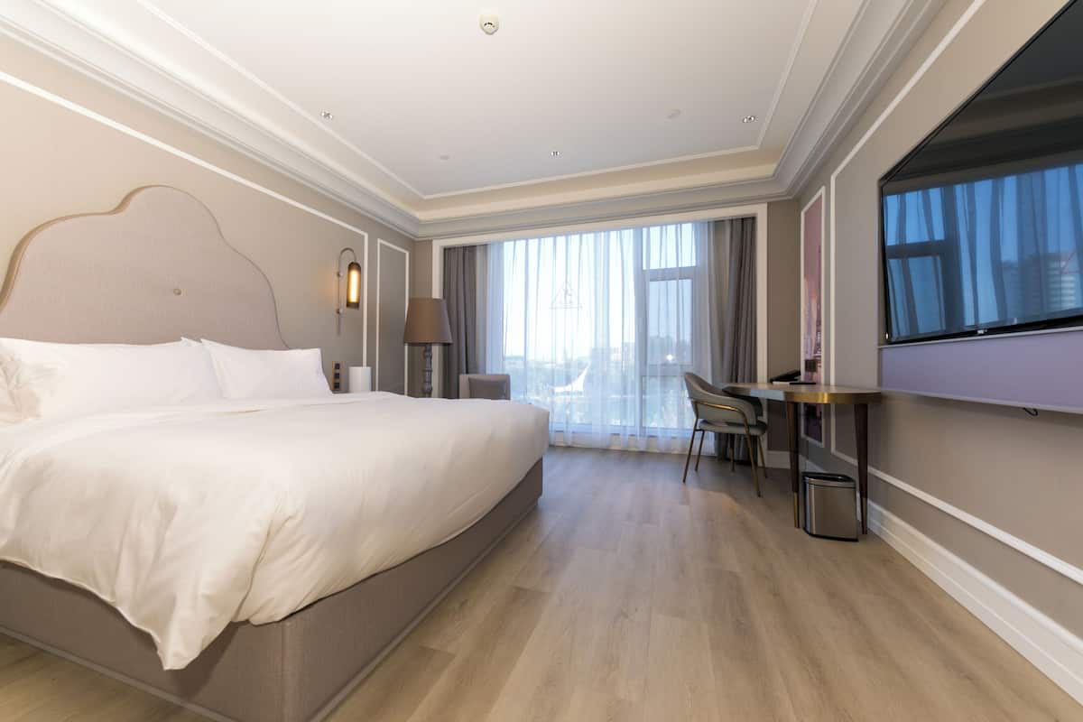 Quarto de hotel com uma cama grande, uma escrivaninha, uma cadeira e vista da cidade através de amplas janelas.