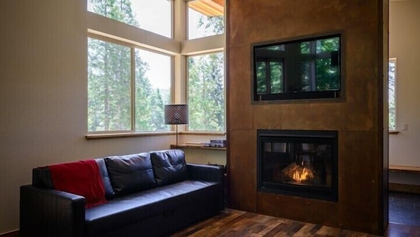 Ein gemütlicher Wohnraum mit Kamin, einer schwarzen Ledersofa und großen Fenstern mit Blick auf Bäume.