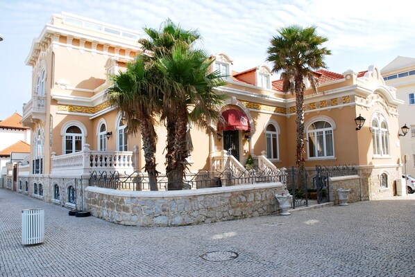 A historic building with a cobblestone courtyard, palm trees, and a red awning.