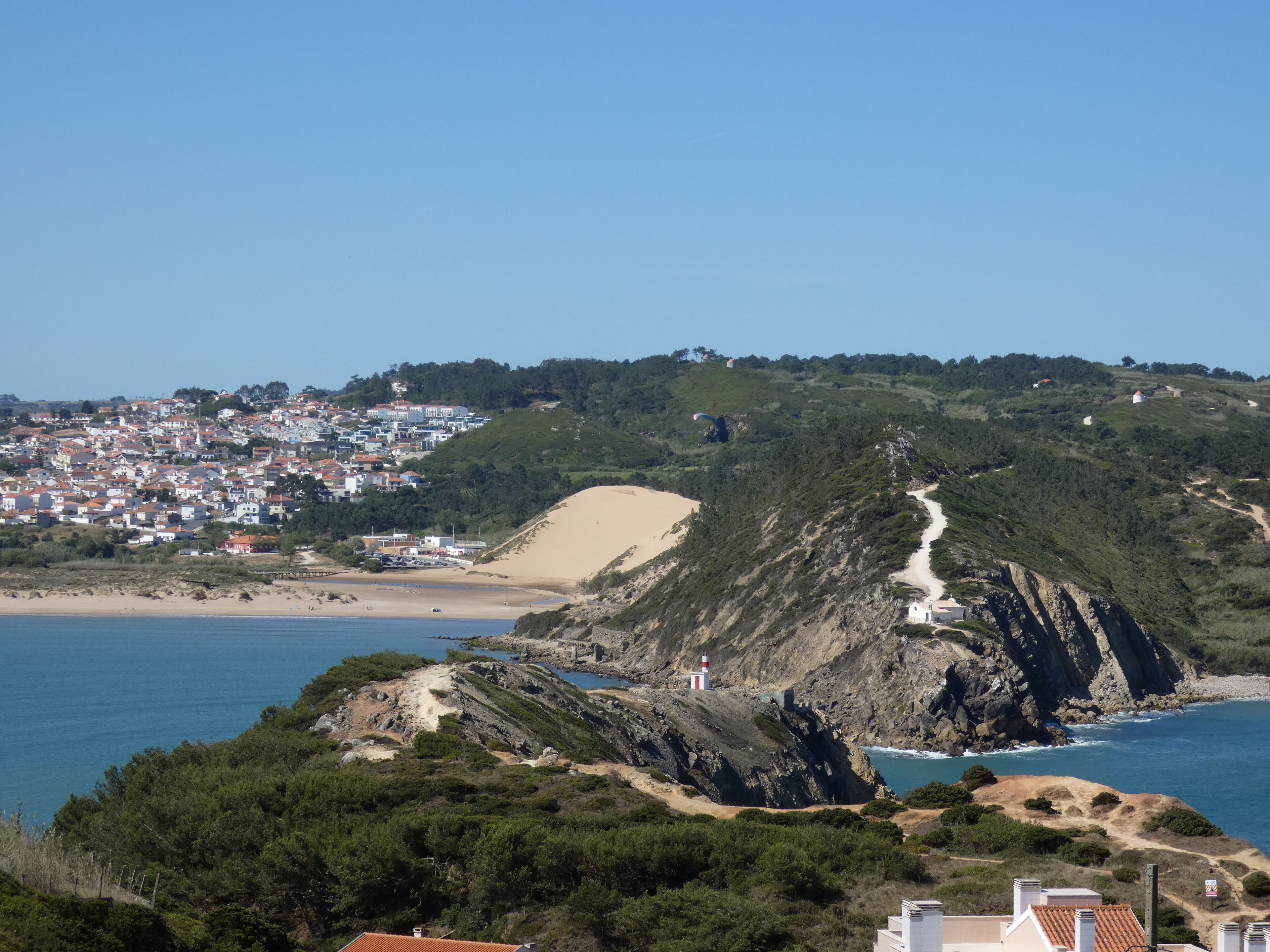 Un pueblo costero con un faro, playa de arena y una carretera sinuosa que sube la colina.