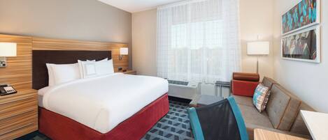 Ein Hotelzimmer mit einem großen Bett, einem Schreibtisch, einem Sessel, einem Nachttisch, einer Lampe und einem Fenster mit durchsichtigen Vorhängen.
