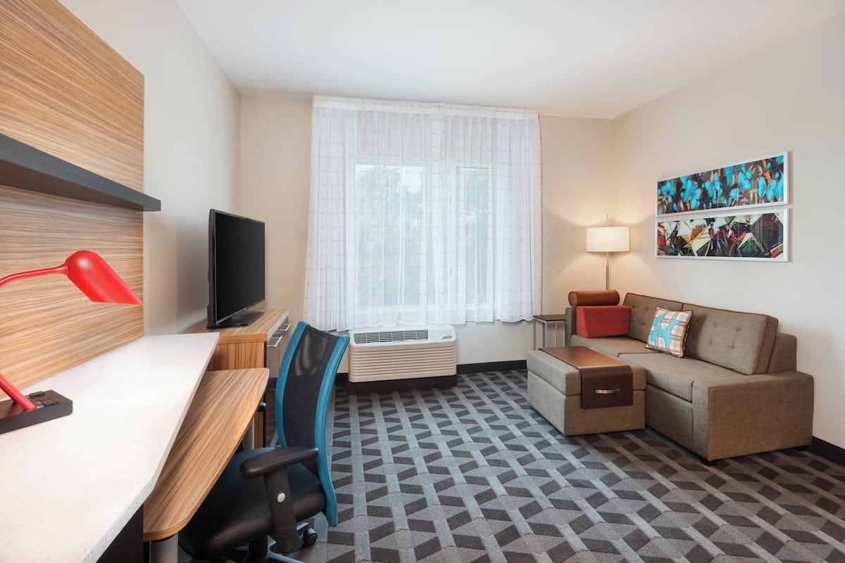 Une chambre d’hôtel équipée d’un bureau, d’une chaise, d’un canapé, d’une télévision, d’un climatiseur, d’une lampe et d’œuvres d’art accrochées au mur.