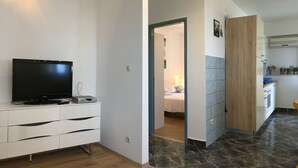 Una sala de estar moderna con un televisor de pantalla plana sobre un mueble blanco, piso de madera y una zona de cocina con un mueble de madera y un lavabo.