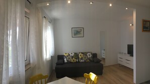 Un salón moderno con un sofá oscuro, una mesa blanca y una silla amarilla. Hay un cuadro en la pared y un televisor sobre un mueble blanco.