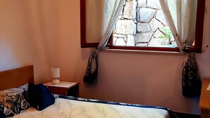 A bedroom with a bed, a nightstand, a lamp, a window with curtains, and a stone wall. - Apartments Cumbelic - One Bedroom Apartment with Balcony and Sea View A3 (Kozarica)