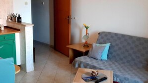A small living area with a sofa, a coffee table, a small table with a vase and a lamp, and a kitchenette with a green cabinet and a countertop. - Apartments Cumbelic - One Bedroom Apartment with Balcony and Sea View A3 (Kozarica)