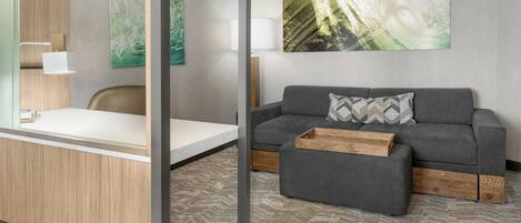A modern hotel room with a grey sofa, a wooden side table, a glass partition, and a large abstract painting on the wall.