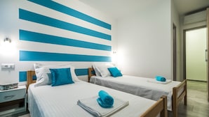 A hotel room with two beds, each with a blue pillow and a rolled towel on the bed. There is a nightstand with a lamp and a small table with a tray. The room has a modern design with blue and white stripes on the wall. - Apartments Sunshine Home - Twin Room (Blue) (Vela Luka)